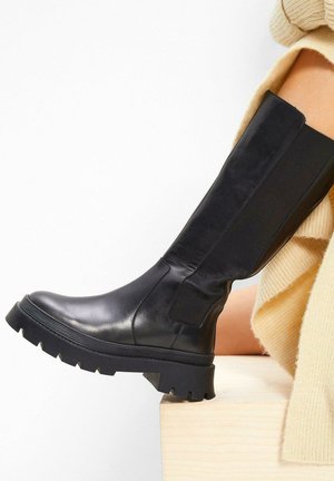 Stivale nero al ginocchio realizzato in pelle liscia con pannello laterale testurizzato. Suola in gomma spessa con un motivo a battistrada per una migliore aderenza.