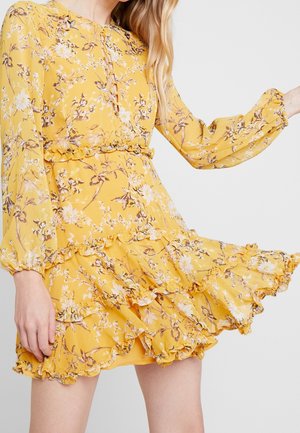 Woman wearing a yellow floral long-sleeve dress with ruffled layers and elastic cuffs, mid-movement against a plain background.