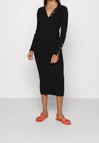 Černé šaty s dlouhým rukávem a ribovaným vzorem, s výstřihem do V a zlatými knoflíky na límci a manžetách, doplněné oranžovými sandály na řemínky.