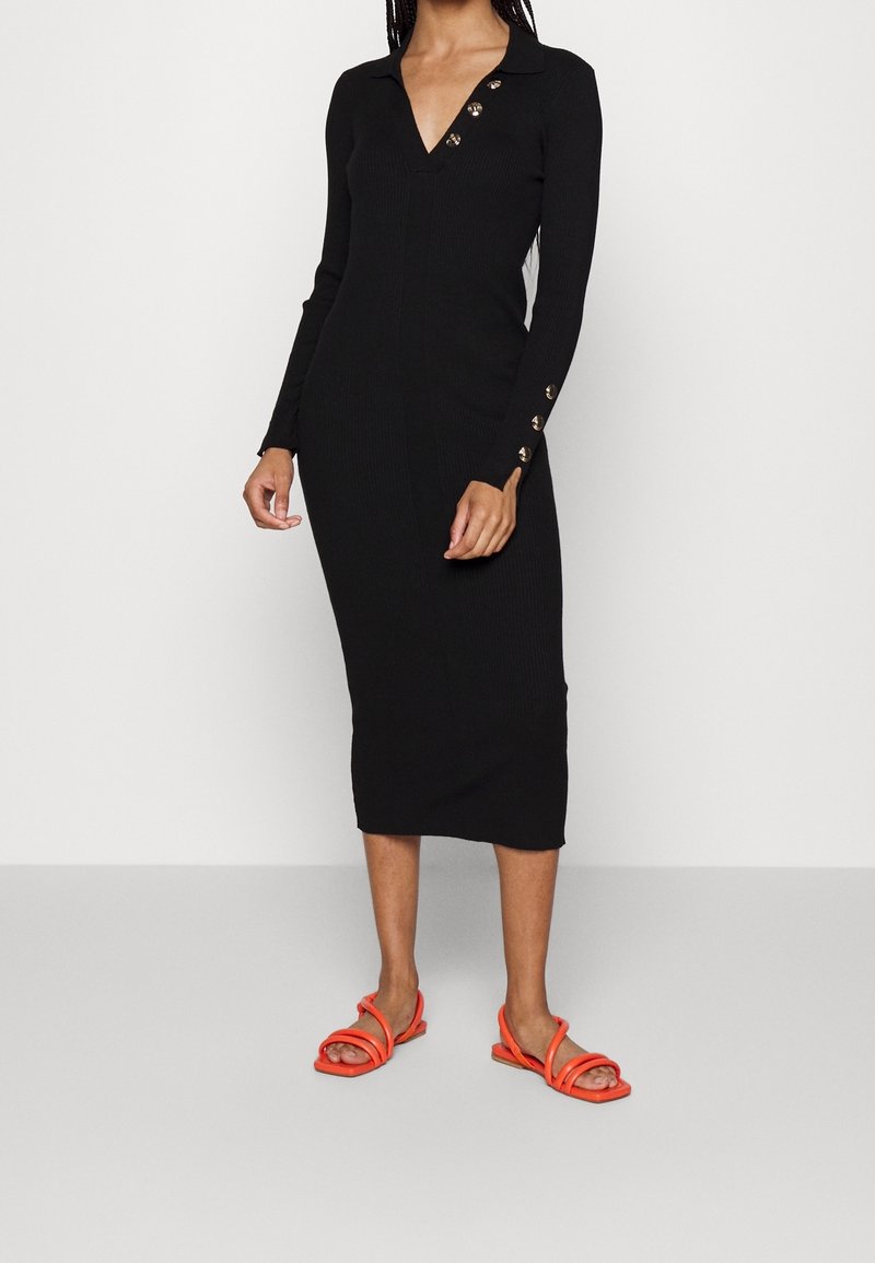 Černé šaty s dlouhým rukávem a ribovaným vzorem, s výstřihem do V a zlatými knoflíky na límci a manžetách, doplněné oranžovými sandály na řemínky.