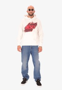 Felpa bianca con grafica rossa e nera "DIESEL", tasca frontale. Abbinata a jeans azzurri chiari e sneakers nere. Indossa occhiali da sole tondi.
