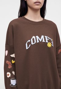Γυναίκα που φοράει καφέ φούτερ με κείμενο "COMFY" και χαμογελαστό πρόσωπο στο μπροστινό μέρος και γραφικά με θέμα το πρωινό στα μανίκια.