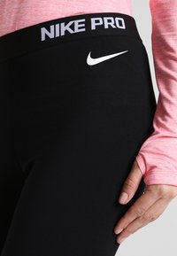 Czarne legginsy Nike Pro z szerokim pasem, na którym widnieje biały napis "NIKE PRO" oraz logo. Materiał wydaje się gładki i elastyczny.