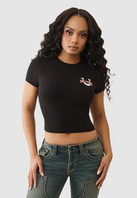 Czarny skrócony t-shirt z miękkiej tkaniny, z małym graficznym logo w kolorze czerwonym i białym po lewej stronie. W zestawie z wzorzystymi niebieskimi dżinsami.