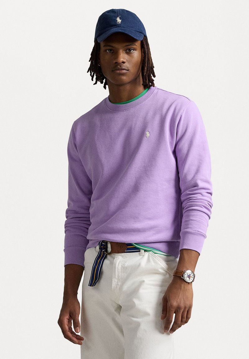 Sweatshirt de algodão roxo com decote redondo e pequeno logotipo; combinado com calças brancas e um cinto listrado multicolorido; boné azul marinho.