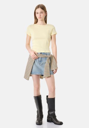 Jeune femme portant un haut jaune pâle à manches courtes, une jupe en jean avec un pull rayé noué à la taille, et des bottes noires montantes, debout sur fond blanc.