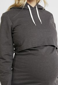 Bluza ciążowa w ciemnoszarym kolorze, o krótkim kroju, z długimi rękawami, przednią kieszenią typu kangurka oraz białymi sznurkami przy kapturze.