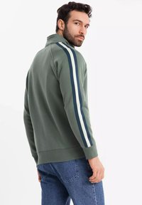 Sudadera verde con cremallera en el cuello y rayas blancas y marinas a lo largo de las mangas. Material suave, puños acanalados y un ajuste casual. Jeans de mezclilla visibles.