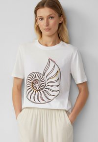 Vrouw met blond haar die een wit T-shirt draagt met een groot bruine spiraal zee-schelp ontwerp met kleurrijke accenten, gecombineerd met lichtbeige broek.