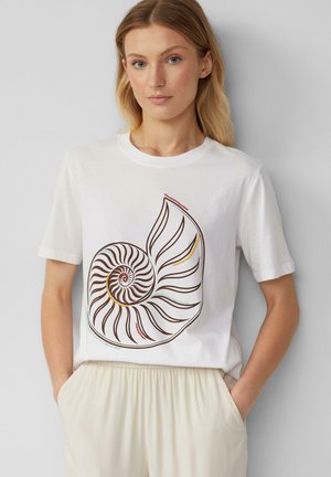 Vrouw met blond haar die een wit T-shirt draagt met een groot bruine spiraal zee-schelp ontwerp met kleurrijke accenten, gecombineerd met lichtbeige broek.