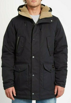 Uomo che indossa una giacca invernale nera con cappuccio foderato in pelliccia sintetica e molte tasche frontali con bottoni e cerniere.
