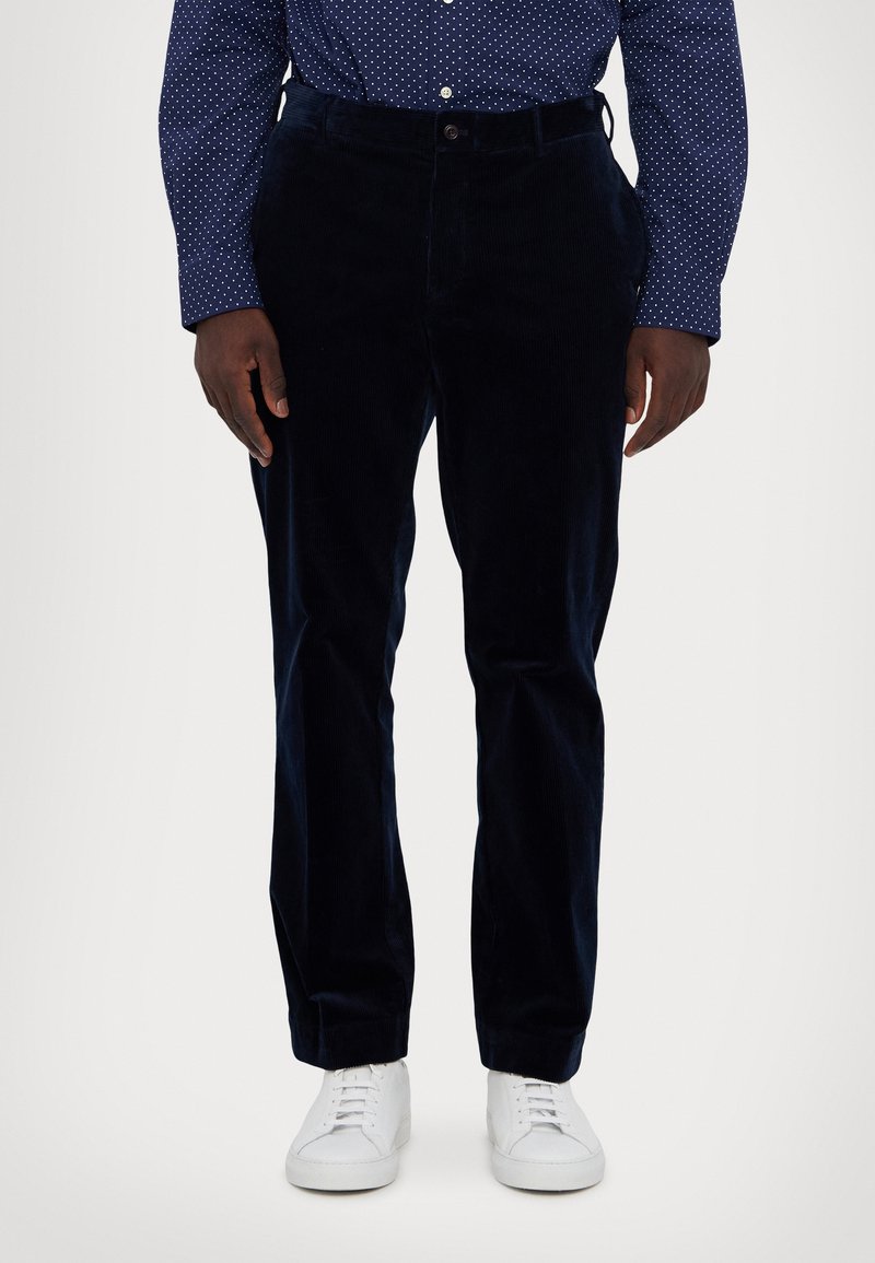 Marineblå kordfløyelsbukser med rett ben-design, med knappefeste og sidelommer, kombinert med hvite sneakers og en prikkete skjorte.