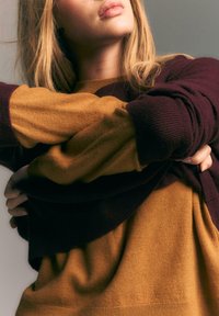 Blondynka w kolorze bordowym i musztardowym swetrze w kolorowe bloki z założonymi na piersiach rękami, stojąca na szarym tle.