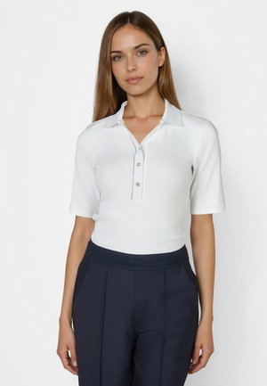 Woman with long brown hair wears a white short-sleeve collared shirt and navy pleated pants, standing against a plain white background.
