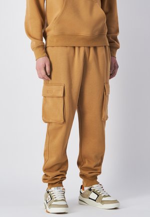 Mężczyzna w musztardowych dresowych spodniach cargo i pasującej bluzie, w białych i beżowych sneakersach, stojący na białym tle.