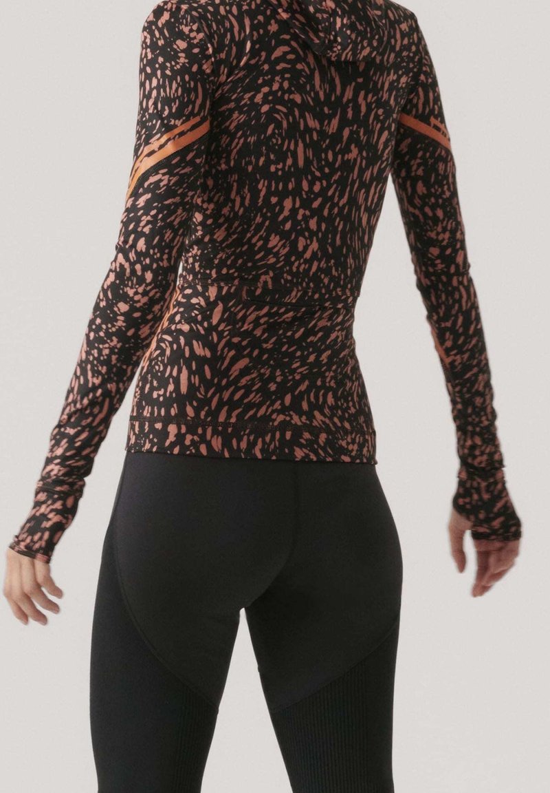 Personne portant un haut de sport à manches longues à motifs avec des trous pour les pouces et des leggings noirs unis, vue de dos sur un fond uni.