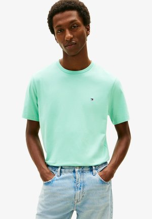 Uomo con le mani nelle tasche che indossa una t-shirt verde menta a collo tondo e jeans azzurri, in piedi su sfondo bianco.