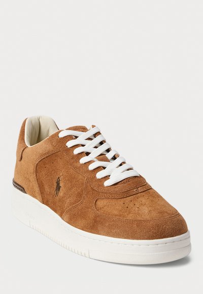 Sneaker in suede marrone con lacci bianchi e suola in gomma bianca. Presenta perforazioni sulla punta e un logo sul lato.