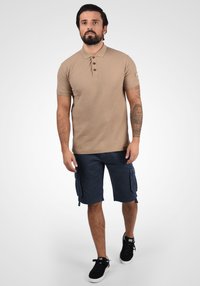 Beige Poloshirt mit drei Holzknöpfen, kurzen Ärmeln; kombiniert mit marineblauen Cargo-Shorts mit seitlichen Taschen und entspannter Passform.