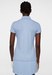 Jasnoniebieska koszulka polo golfowa z teksturowanym wzorem, z klasycznym kołnierzykiem i krótkimi rękawami. Biały detal z logo na plecach w okolicy szyi.