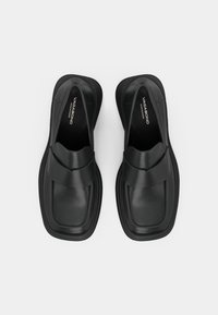Black slip-on shoes with a smooth leather finish, rounded toe, thick sole, and a prominent strap across the upper. Minimalist design.