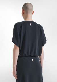 Persoon met geschoren hoofd, gekleed in een loszittend zwart topje met korte mouwen en een bijpassende rok, gezien van achteren tegen een witte achtergrond.