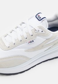 Sneaker bianca con dettagli in suede beige, caratterizzata da un design testurizzato, lacci bianchi piatti e un logo blu e rosso sulla linguetta.