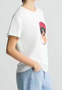 Weißes Baumwoll-T-Shirt mit kurzen Umschlagärmeln, das einen bunten Grafikdruck einer Frau vor rotem Hintergrund zeigt.