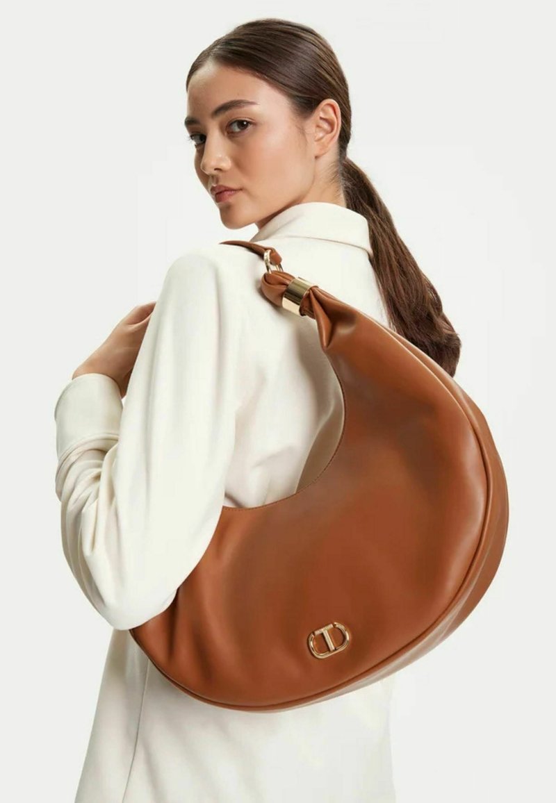 Borsa a spalla in pelle marrone a forma di mezzaluna, con accessori in oro e una texture morbida, indossata sopra una camicia bianca.