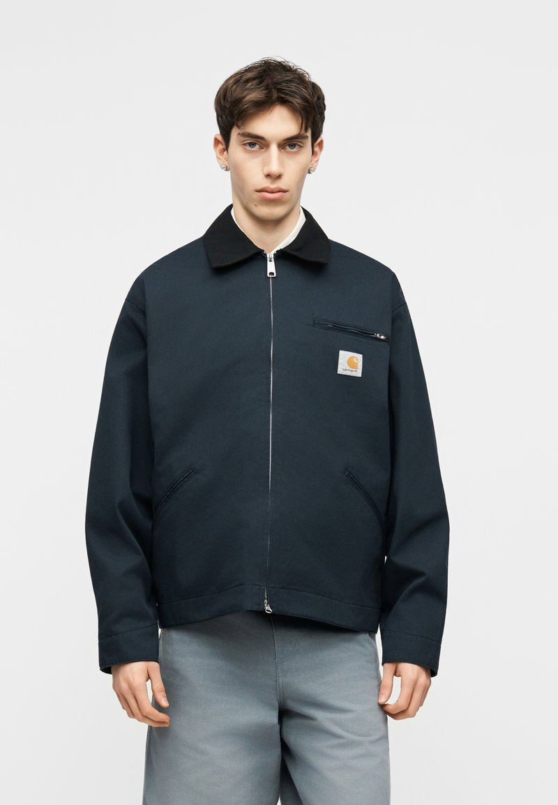 Jonge man met kort donker haar, gekleed in een zwart jack met ritssluiting en een Carhartt-logo op de borst, en een lichtgrijze broek, staand tegen een witte achtergrond.