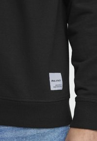 Zwarte sweatshirt met ribgebreide manchetten en onderrand, voorzien van een klein wit label met zwarte tekst. Soepele textuur en eenvoudig ontwerp.
