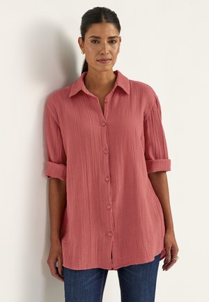 Kvinde med mørkt hår i en lav hestehale iført en tekstureret koralfarvet skjorte med knapper og ærmer rullet op til albuerne samt blå jeans.