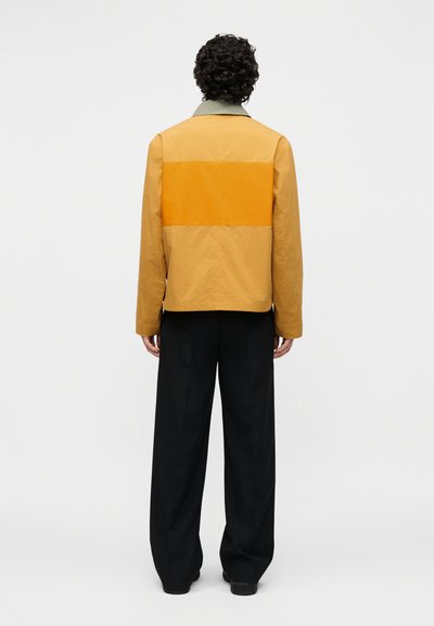 Άτομο με σγουρά μαλλιά που στέκεται με την πλάτη, φοράει μια μπεζ και πορτοκαλί μπουφάν με χρωματικούς τομείς και φαρδιά μαύρη παντελόνια σε λευκό φόντο.