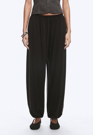 Femme portant un pantalon noir ample avec des poignets élastiques, associé à des chaussures noires à enfiler et un haut court sombre sans manches.