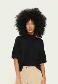 Pulover scurt din tricot ribatat, de culoare neagră, cu mâneci scurte și guler înalt, asortat cu pantaloni din velur bej, care dispun de buzunare frontale și închidere cu un singur buton.