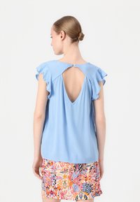 Blusa azzurra a maniche corte con bordi arricciati e un dettaglio a buco della serratura sul retro, abbinata a una gonna con fantasia colorata. Il tessuto sembra morbido.