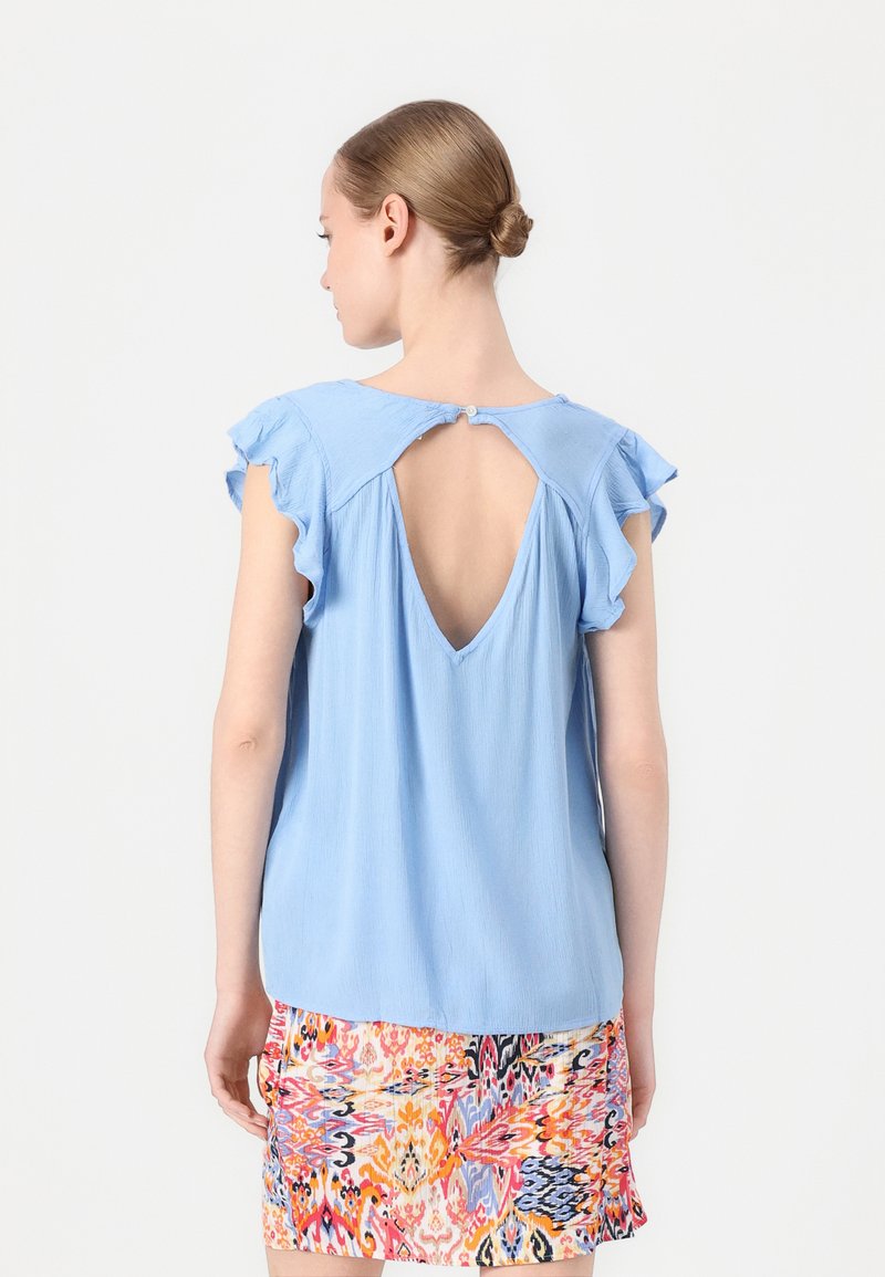 Blusa azzurra a maniche corte con bordi arricciati e un dettaglio a buco della serratura sul retro, abbinata a una gonna con fantasia colorata. Il tessuto sembra morbido.