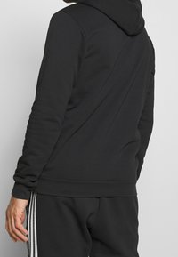 Svart hoodie med en texturerad finish, med en rymlig huva och åtsittande ärmslut, visad tillsammans med matchande mörka sweatpants.