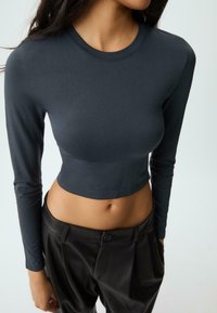 Frau in einem figurbetonten, langärmeligen, dunkelgrauen Cropped-Top, kombiniert mit hochgeschnittenen, lockeren schwarzen Hosen vor einem schlichten Hintergrund.