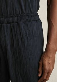 Marineblauwe textuurbroek met een golfpatroon, voorzien van een elastische tailleband en een zijzak. Close-up van de stof en een hand.