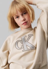 Sudadera beige con escote redondeado y mangas largas. Presenta un gran logo "CK" plateado y brillante en la parte delantera. Textura suave.