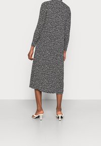 Mujer vestida con un vestido midi negro con estampado floral blanco y mangas largas, combinado con sandalias de tacón blancas, de pie frente a un fondo liso.