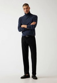 Pull col roulé marine, texture douce, coupe ajustée, associé à un pantalon noir droit et des mocassins noirs. Le mannequin est debout, les bras croisés.