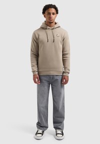 Man staat gekleed in een beige hoodie, losse grijze jeans en zwart-witte sneakers tegen een effen achtergrond.