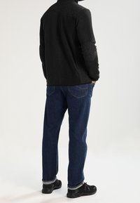 Schwarze Fleecejacke mit grauen Ellbogenflicken, kombiniert mit blauen Denim-Jeans. Schwarze Sneaker mit strukturierten Oberflächen vervollständigen das Outfit.