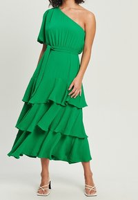 Zelené maxi šaty na jedno rameno s tiered volány, texturovanou látkou, úzkým pasem a splývavým střihem. Nosené s bílými sandály na šněrování.