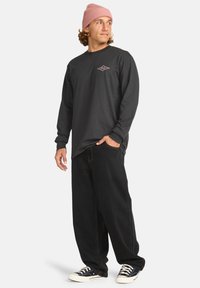 Schwarz langärmliges Shirt mit einem diamantförmigen Logo auf der Vorderseite, kombiniert mit lockeren schwarzen Hosen und schwarzen Sneakers. Das Model trägt eine hellrosa Mütze.