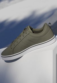 Zapatilla baja de color verde oliva con suela blanca y cordones, apoyada sobre una superficie blanca con fondo sombreado.