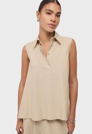 Femme portant une chemise beige sans manches avec col et boutons, boucles d'oreilles argentées en forme d'anneau et un bracelet, debout devant un fond clair uni.