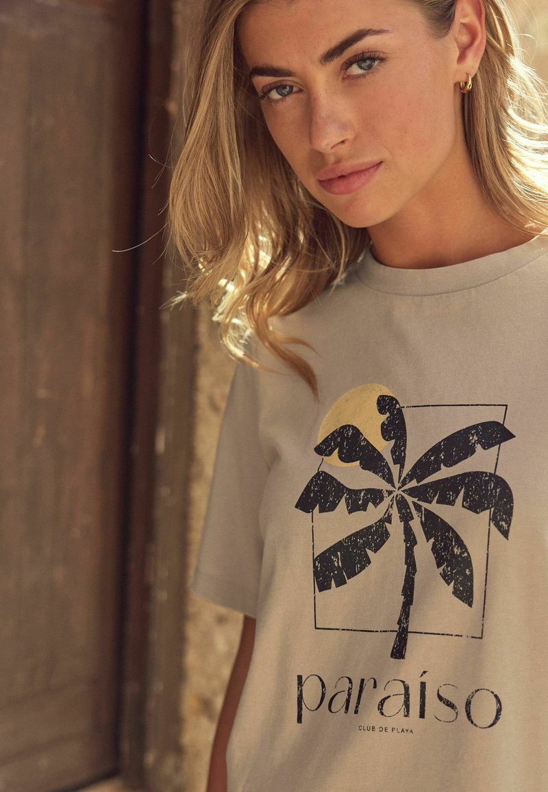 Grijze katoenen t-shirt met een grote palmboomafbeelding in het zwart en een gele zon, met de tekst "paraiso" en "club de playa" eronder.